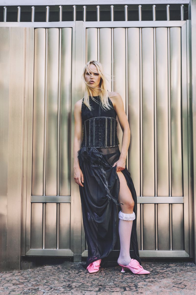 A blonde-haired fashion model poses against a silver metal door wearing a black top and skirt, pink heels, and white socks.