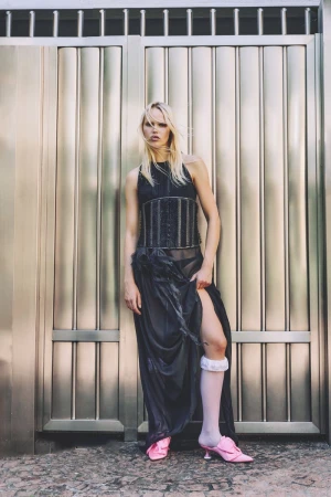 A blonde-haired fashion model poses against a silver metal door wearing a black top and skirt, pink heels, and white socks.