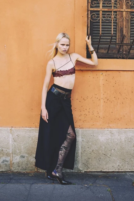 Fashion model poses in burgundy bra top, black skirt, and floral tights against an orange wall near a window with metal bars.