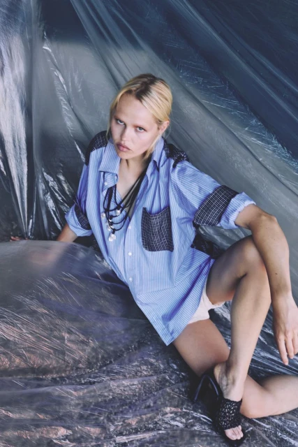 Fashionable fair-skinned woman with blonde hair poses in an unbuttoned blue striped shirt and black heeled shoes against a blue background.