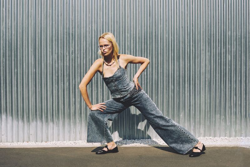 Woman with blonde hair poses in a denim outfit with black shoes in front of a corrugated metal wall in an outdoor fashion shot.