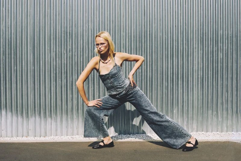 Woman with blonde hair poses in a denim outfit with black shoes in front of a corrugated metal wall in an outdoor fashion shot.