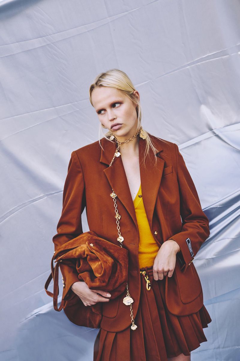 A fair-skinned adult woman poses in a brown blazer and pleated skirt, holding a matching velvet bag, set against a silver backdrop.