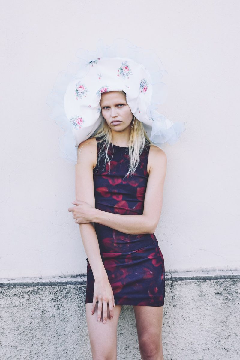A model in a black and red floral dress poses against a wall with a large, white floral headpiece in a fashion editorial.
