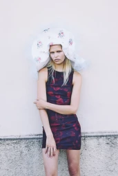 Model wearing a patterned dress and a large floral hat standing against a light wall for Spaghetti Magazine.