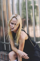 Blonde female model in a black top posing against a vertical metallic ribbed column exterior