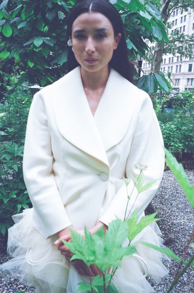 Woman in a cream-colored coat poses in a lush green park with trees and foliage. Natural light illuminates the scene.