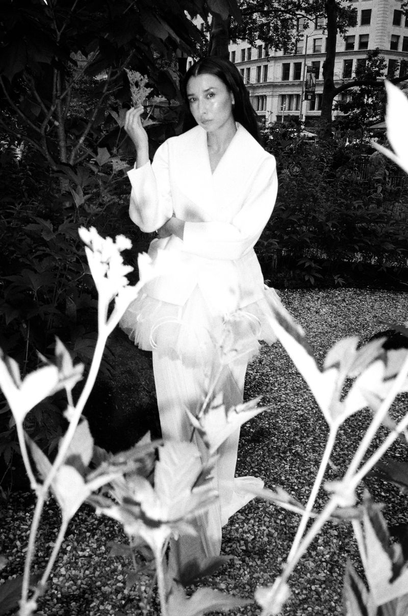 Monochrome portrait of an adult woman in a white dress amid lush green foliage, bathed in soft, diffused natural light in an urban garden.