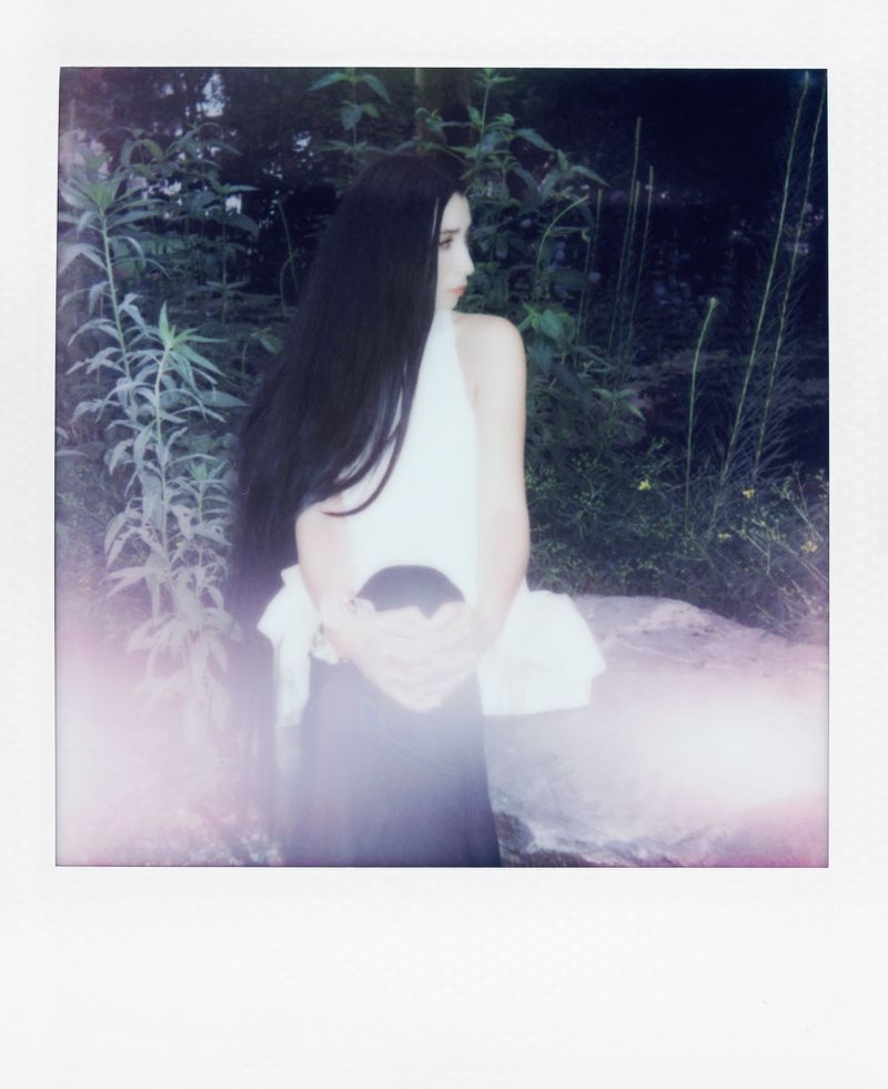 A woman with long dark hair poses in a white top and dark skirt in a natural outdoor setting with greenery in a slightly blurred, dreamy photograph.