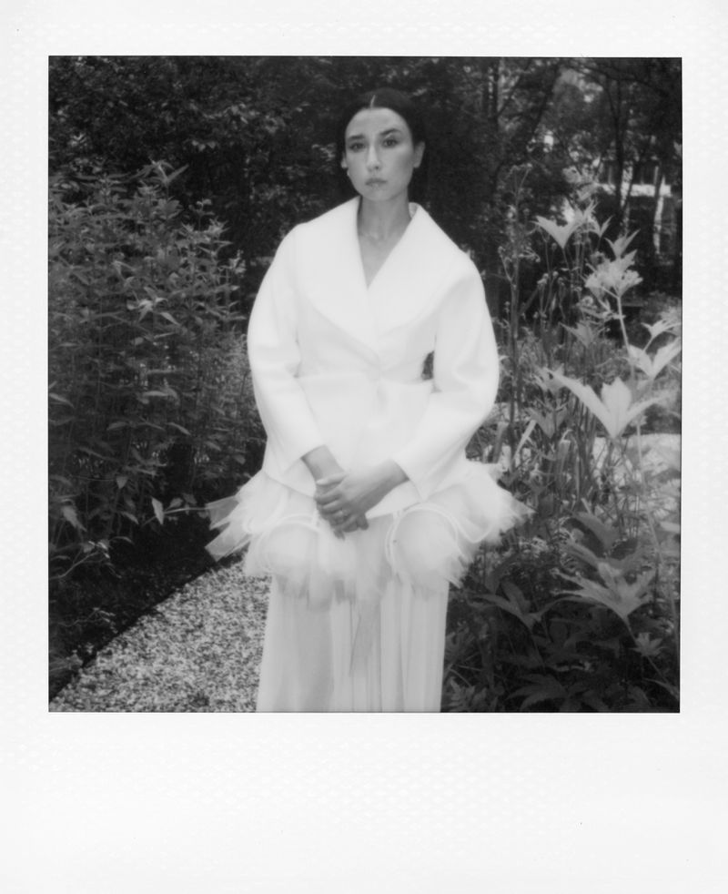 Black and white portrait of a woman in a white dress and jacket, standing outside in a garden with lush greenery and a stone path.