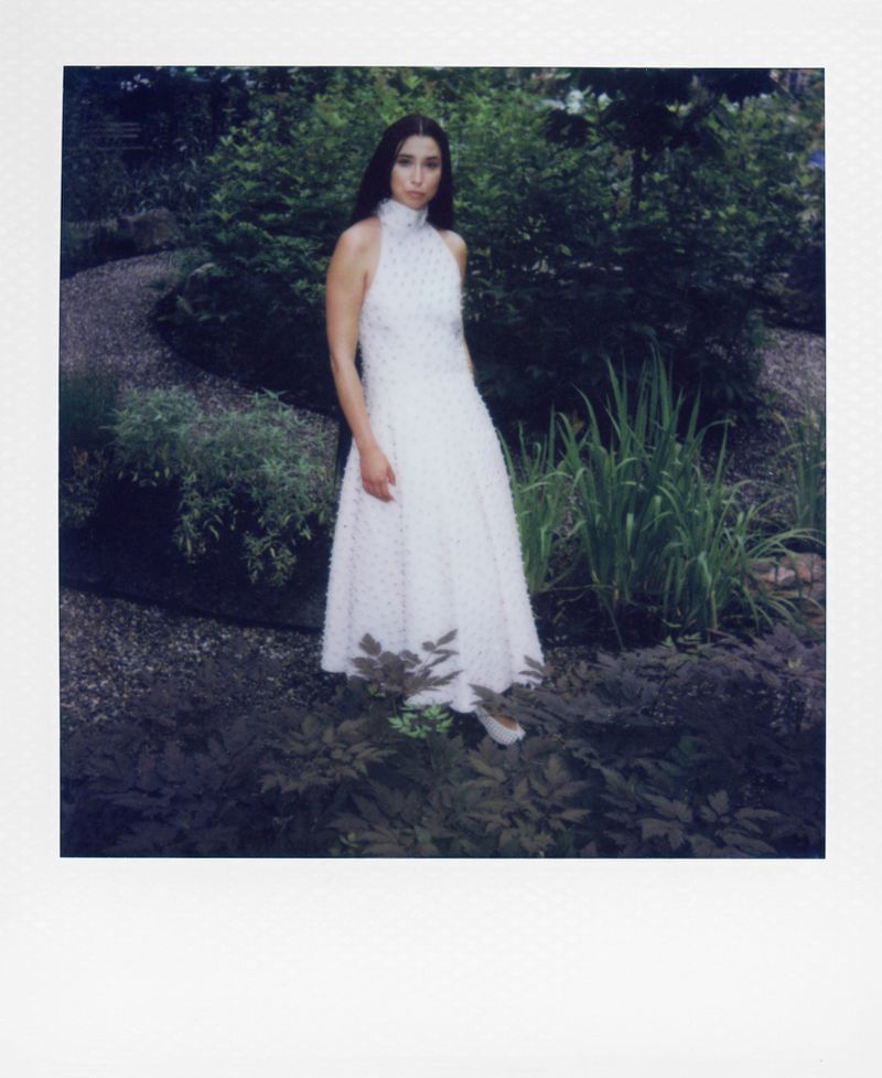 Young woman in elegant white dress poses in garden with greenery and gravel path, captured in a full shot with soft natural light.