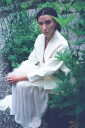 Lily Kwong seated amidst lush green garden foliage wearing an off-white structured jacket and layered skirt for Cero Magazine.