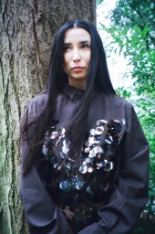 Portrait of Lily Kwong standing outdoors in front of a tree trunk wearing a dark button-down shirt embellished with large reflective discs
