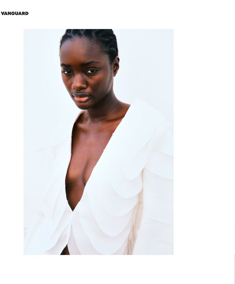 Close-up portrait of a dark-skinned woman with braided hair wearing a white top with draped detailing against a white background.