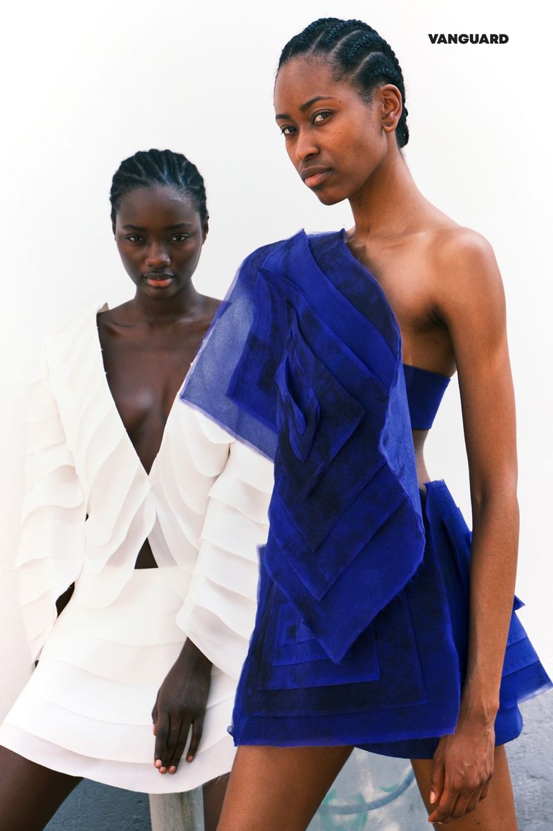 Two African-descent women pose in fashionable attire against a stark background, one in a white ensemble, the other in draped blue fabric.