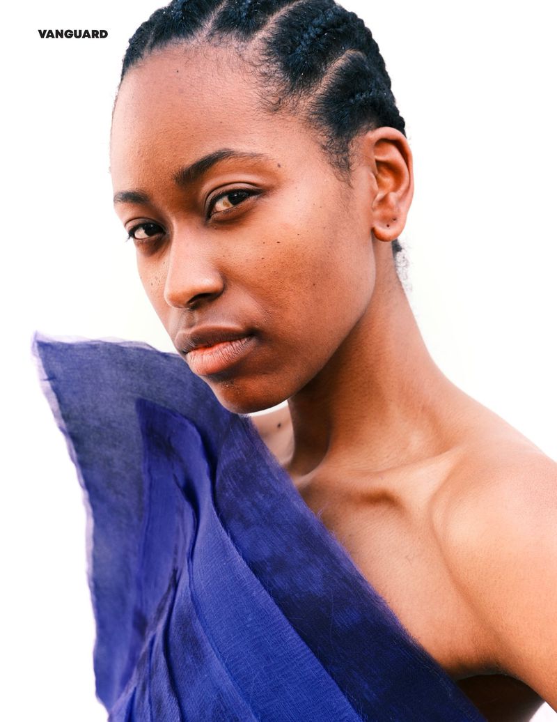 Close-up portrait of a young Black woman with cornrow braids, draped in royal blue fabric against a bright white background in natural light.