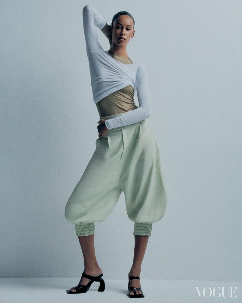 A striking fashion shot features a model in a white top, unique light green pants, and black heels, set against a light backdrop.