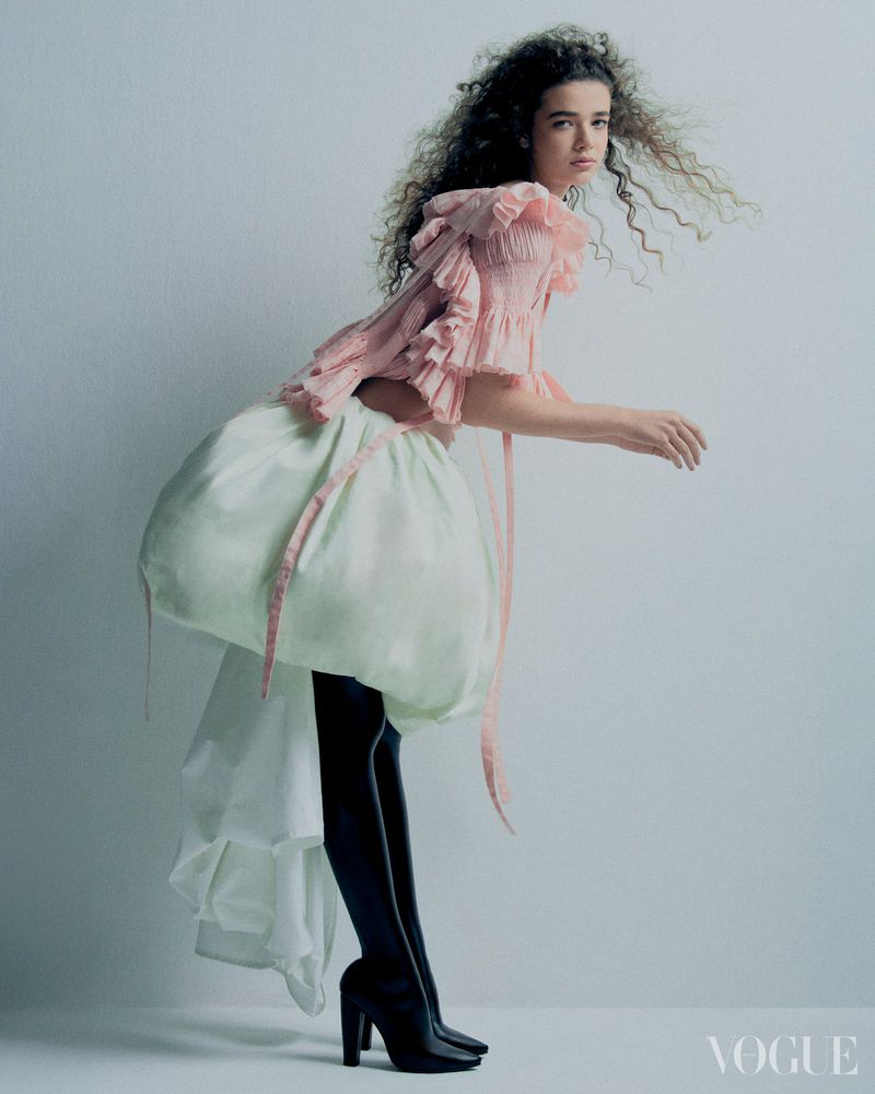 A female model with curly hair poses in a pink ruffled top, black tights, and black platform heels against a clean white background.