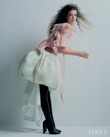 A female model with curly hair poses in a pink ruffled top, black tights, and black platform heels against a clean white background.