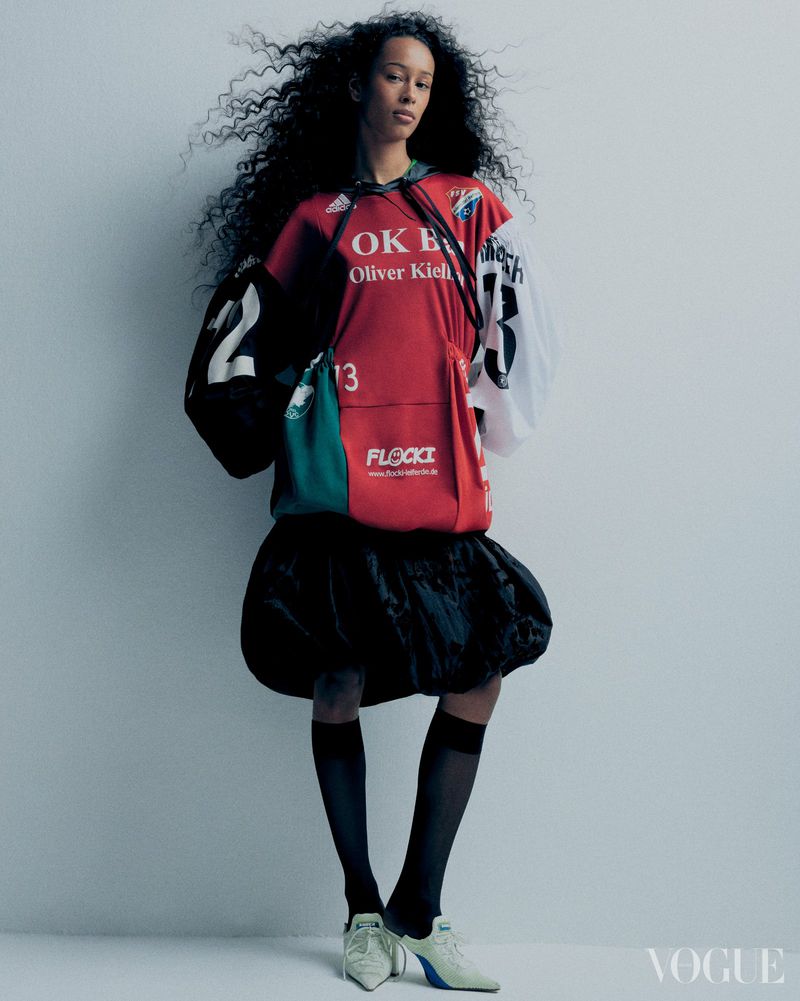 Fashionable adult female in an oversized hoodie and black bubble skirt posing in a photo studio against a solid grey wall.