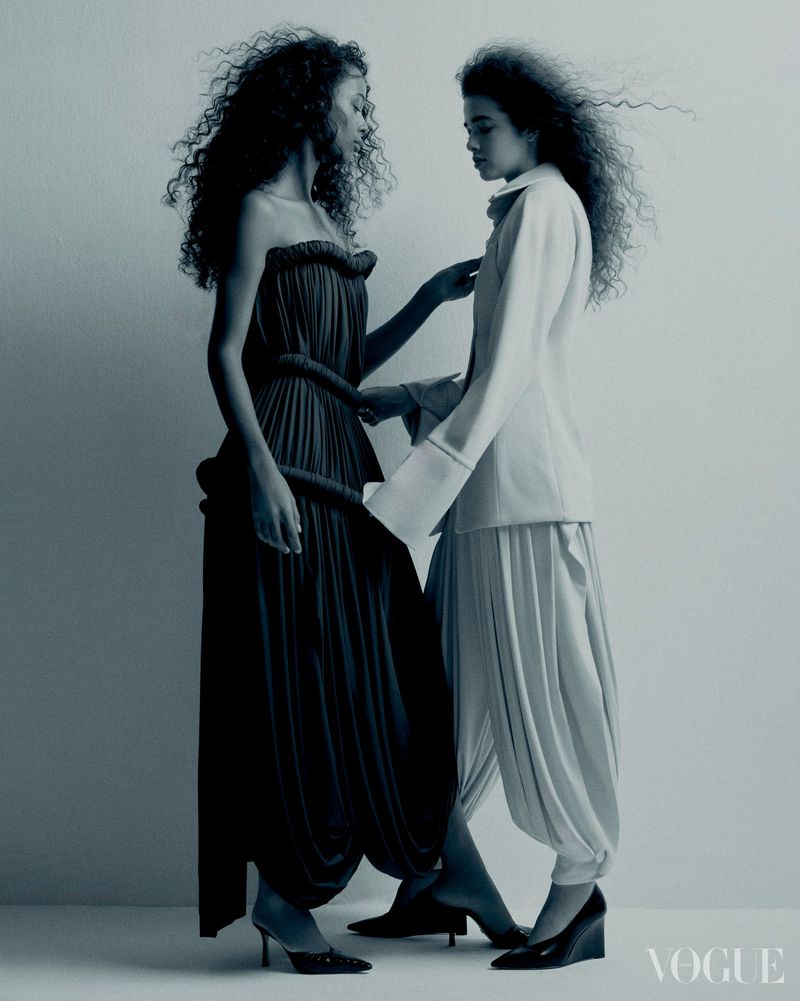 Two fashion models wearing draped, high-fashion clothing in a studio setting. One model wears a dark dress, the other a light colored jacket and pants.