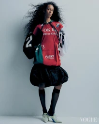Model with voluminous dark curly hair wearing a colour-blocked athletic top and black bubble skirt against a grey background.