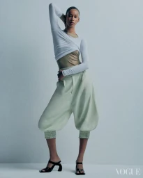Model posing in layered white long-sleeve top, sage green trousers, and black heeled sandals against a plain grey studio backdrop.