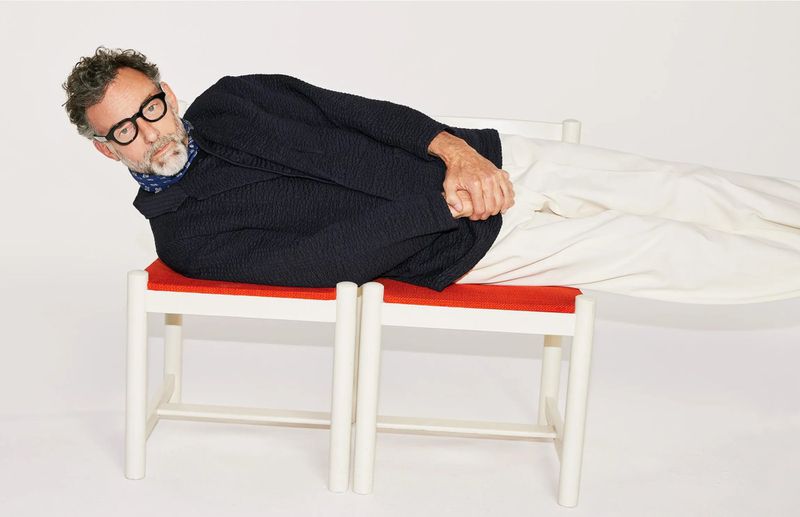 Adult male with beard and eyeglasses lying on two red and white stools in a light studio setting, wearing a patterned navy jacket and white pants.