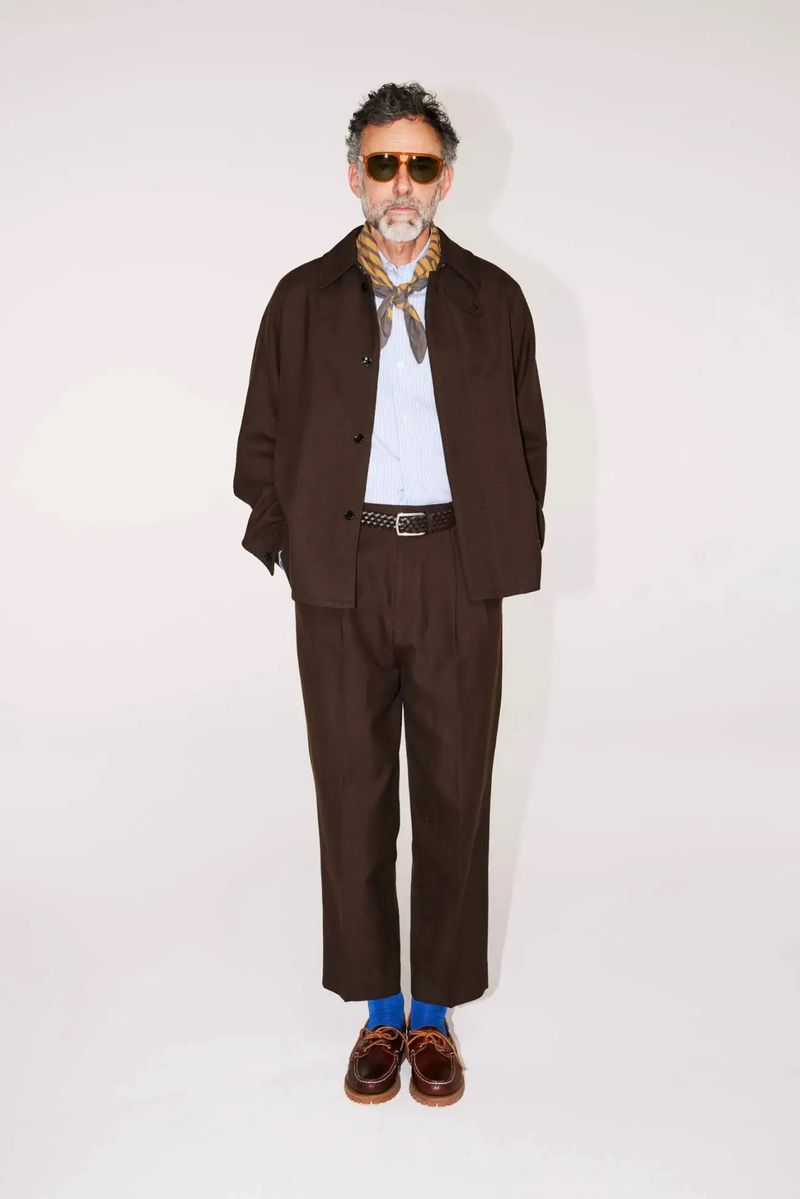 Full-length portrait of a man in a brown suit, blue socks, and brown loafers, wearing sunglasses and a patterned scarf, against a white backdrop.