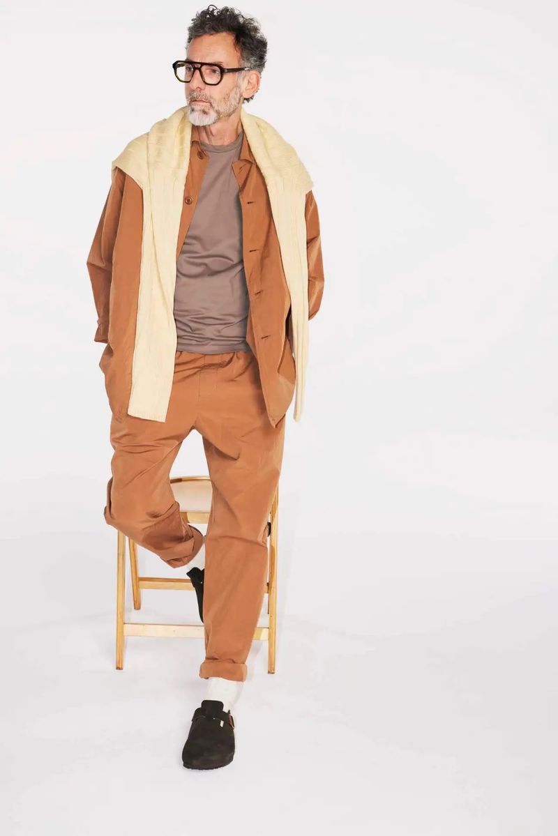 Older adult man in casual attire, eyeglasses, leaning on wooden chair against a plain white backdrop in a brightly lit studio setting.