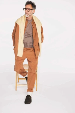 Older adult man in casual attire, eyeglasses, leaning on wooden chair against a plain white backdrop in a brightly lit studio setting.