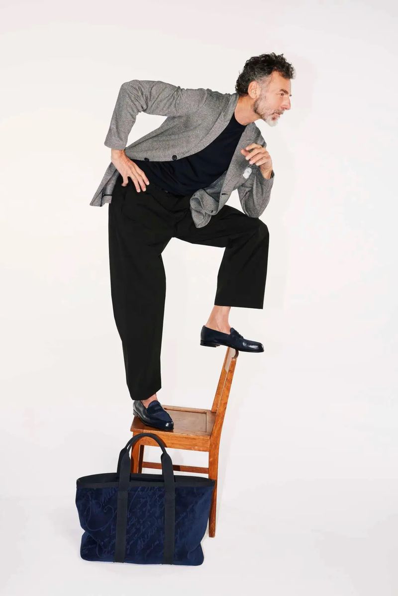 Mature adult man wearing a grey blazer and black slacks stands on a wooden chair with a navy blue patterned tote bag nearby.