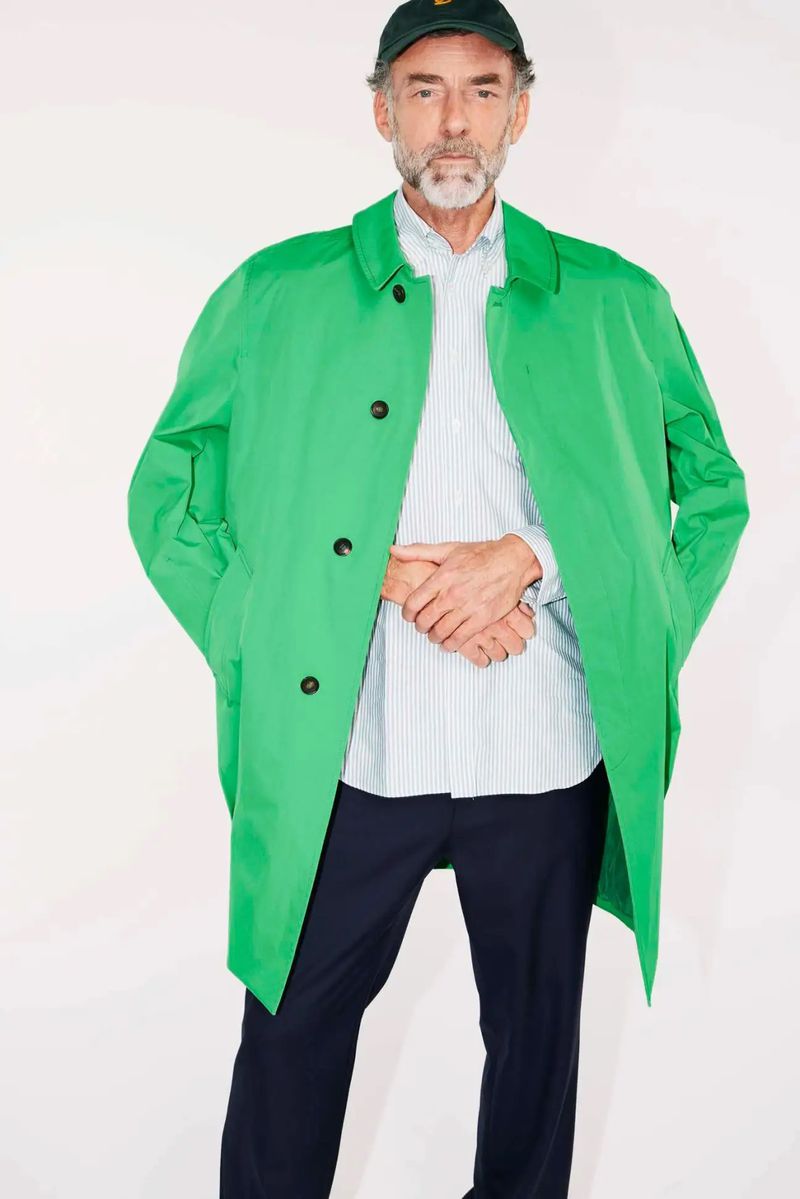 Mature man in green coat and cap stands against white backdrop. Fashion shot of adult male with a salt and pepper beard.