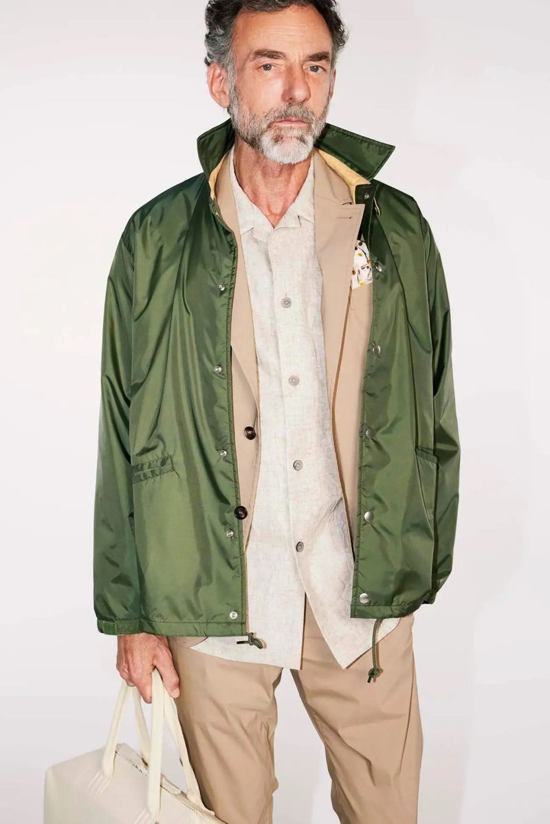 Mature adult male poses in beige pants and layered top, carrying a bag. He wears a short green jacket, against a white backdrop.