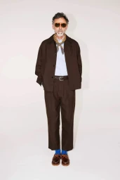 Man wearing a dark brown suit jacket and matching trousers with tinted sunglasses and a patterned neck scarf against a white background
