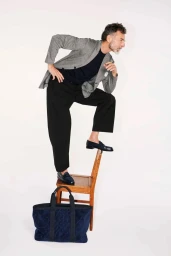 A man standing with one foot on a wooden chair next to a large dark tote bag against a white background for Monocle Magazine.
