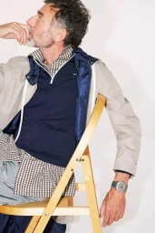 Mature man seated on a wooden chair wearing a navy blue half zip top, checkered shirt, and light jacket against a white background.