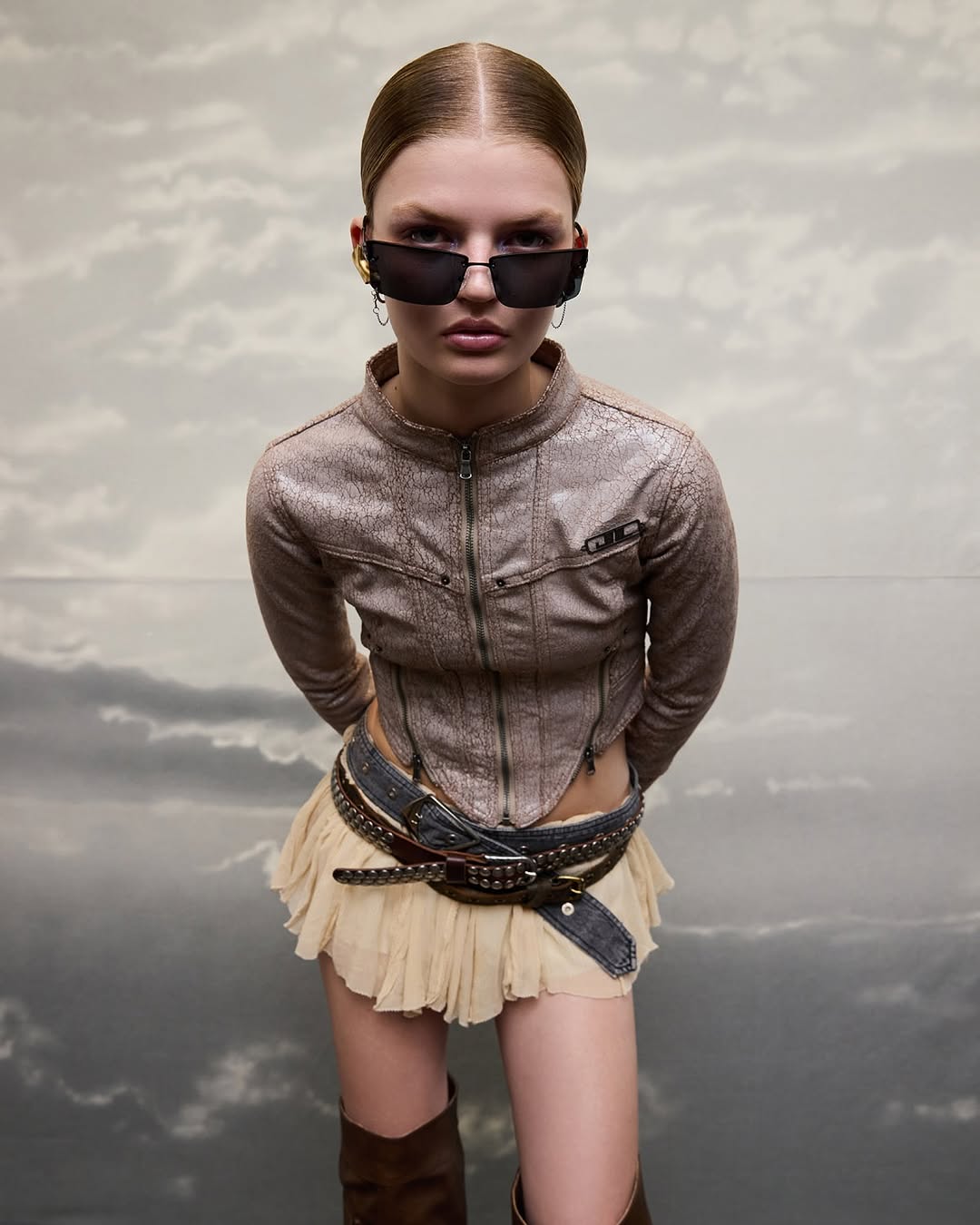Woman poses in a brown jacket, tan mini skirt, and brown boots against a cloudy sky backdrop, accessorized with black sunglasses.