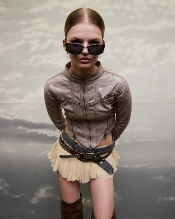 Woman poses in a brown jacket, tan mini skirt, and brown boots against a cloudy sky backdrop, accessorized with black sunglasses.