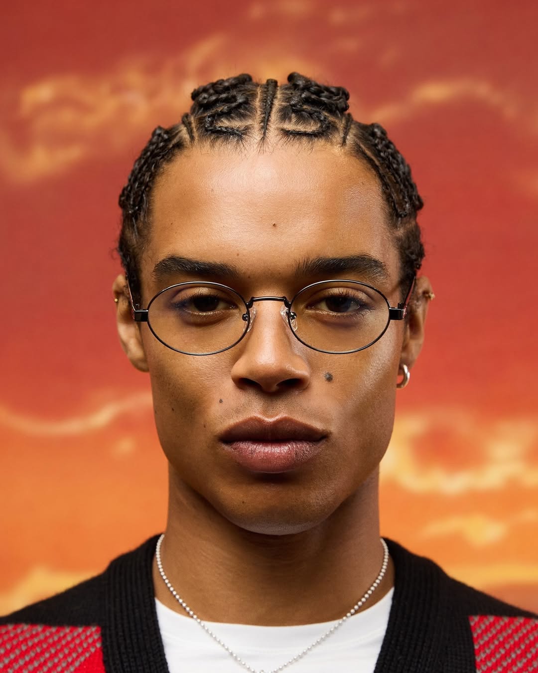 Close-up portrait of a young adult with braided hair wearing oval eyeglasses and a silver chain necklace against a warm background.