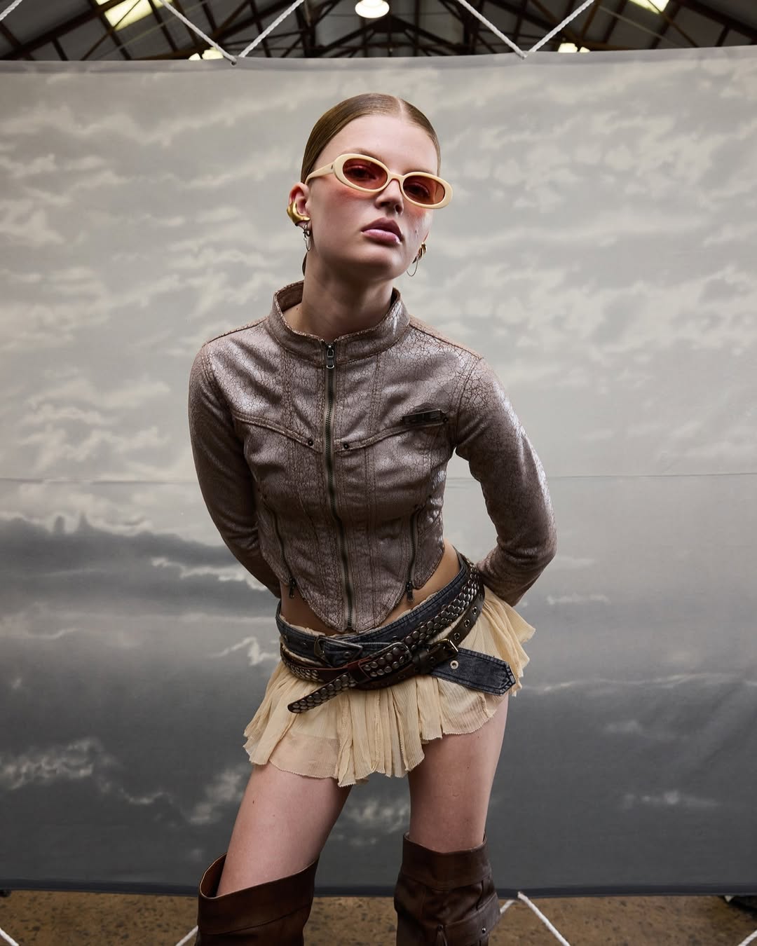 A fair-skinned young woman poses in a silver jacket, tan miniskirt, and brown boots against a cloudy background, wearing sunglasses.