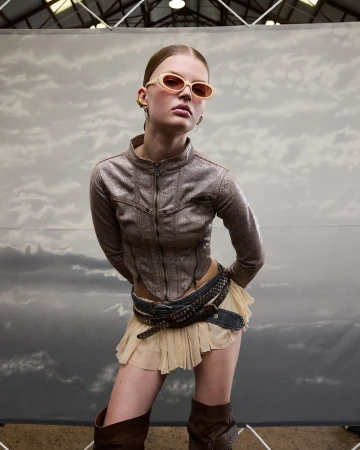 A fair-skinned young woman poses in a silver jacket, tan miniskirt, and brown boots against a cloudy background, wearing sunglasses.