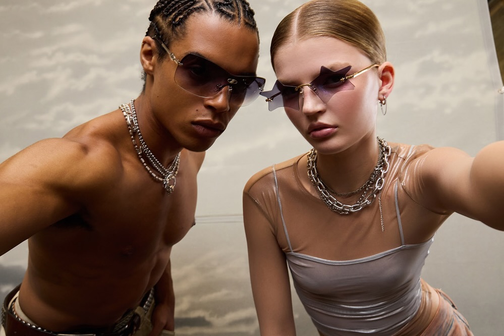 Two fashion models wearing sunglasses and silver necklaces pose in front of an abstract background for a beauty and fashion shoot.