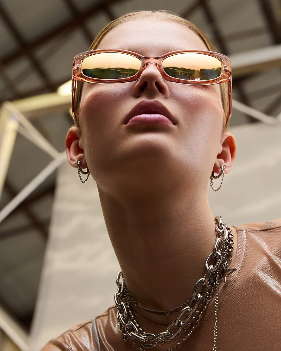 Close-up, low-angle shot of a young adult woman wearing rose-tinted sunglasses and layered metal chain necklaces, earrings, and shiny shirt.