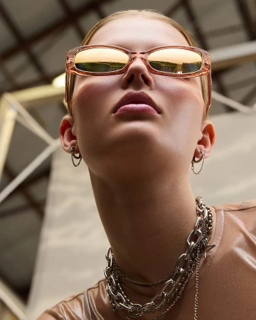 Close-up, low-angle shot of a young adult woman wearing rose-tinted sunglasses and layered metal chain necklaces, earrings, and shiny shirt.