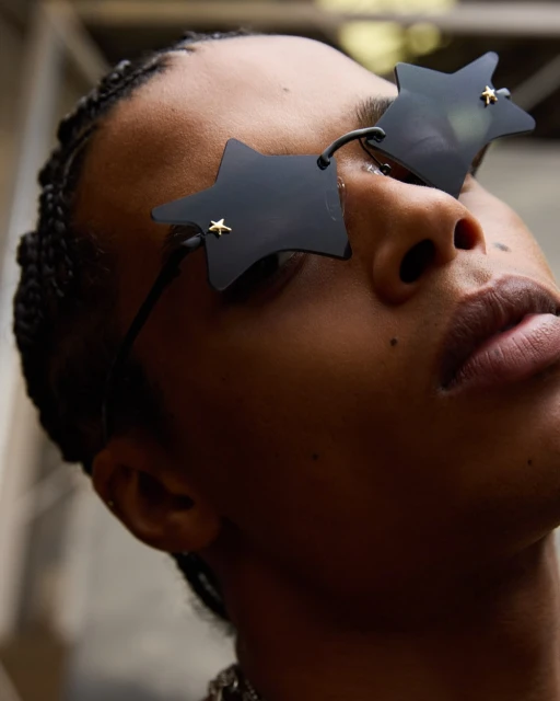 Portrait of a person wearing unique star-shaped sunglasses, shot from a low angle against an indistinct gray background with soft lighting.