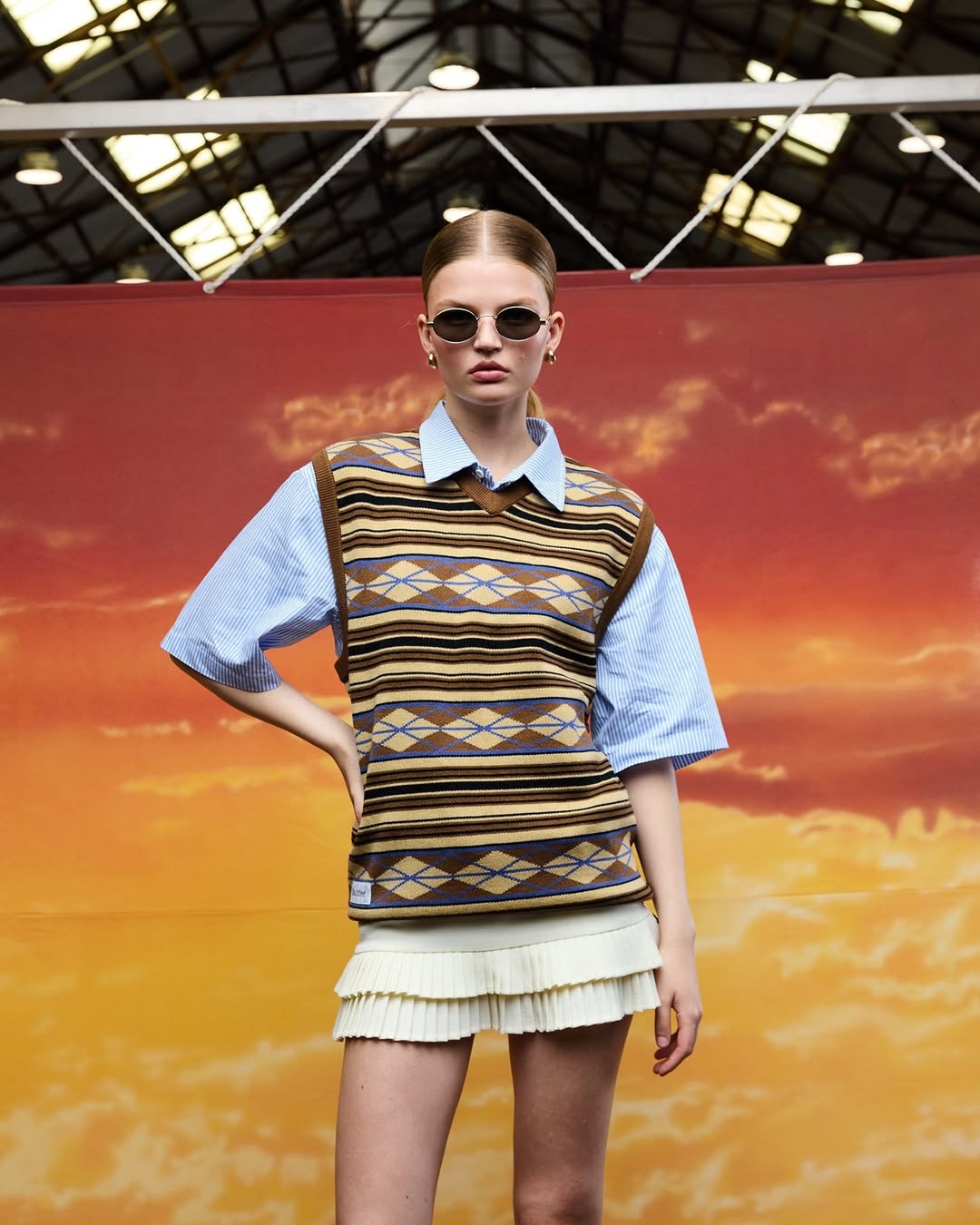 Adult female fashion model posing in a light blue short sleeve shirt, a brown patterned sweater vest, and a white mini skirt against a sky backdrop.