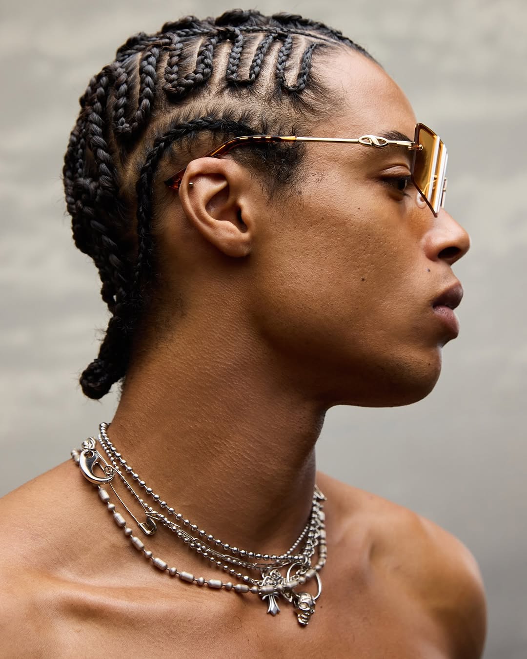 Profile shot of an adult male with braided hair wearing square sunglasses and layered silver necklaces against a neutral background.