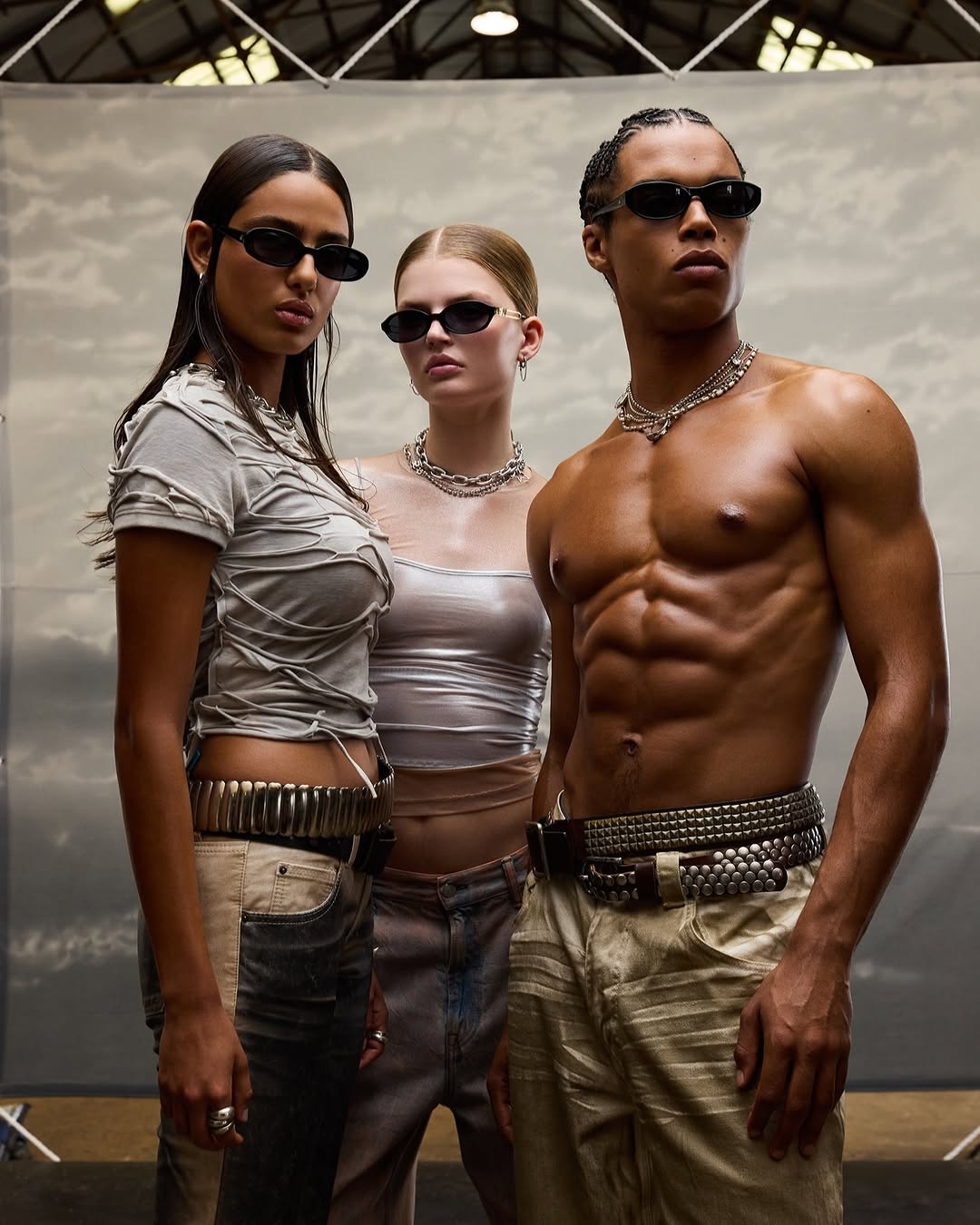Three fashion models wearing stylish clothing and sunglasses pose in a studio with a backdrop, showcasing modern fashion trends.