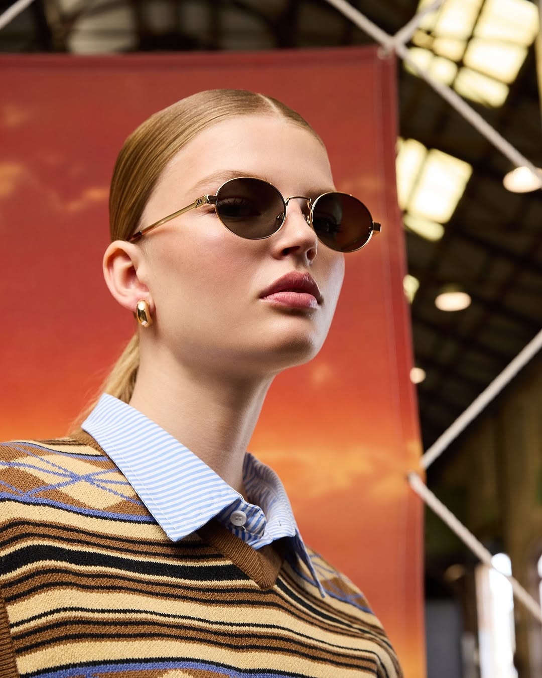 A fair-skinned adult woman with blond hair and round sunglasses poses indoors against a warm-toned backdrop wearing a striped sweater.